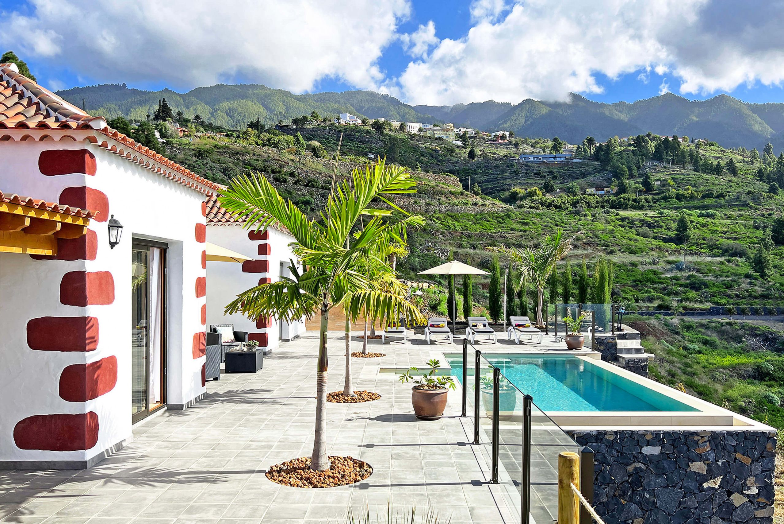 A poolside area with a view of mountains, a white building with red accents, and outdoor seating.
