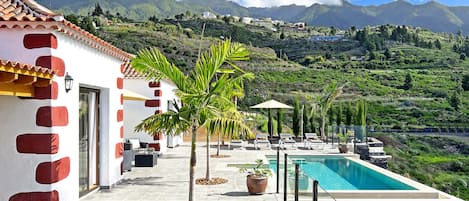 Un espace aménagé au bord de la piscine, avec vue sur les montagnes, un bâtiment blanc agrémenté de touches de rouge et des sièges extérieurs.