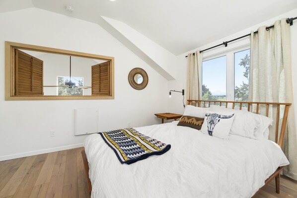A bedroom with a large bed, a window with curtains, a mirror, a bedside table, and a radiator. - Smith Rock Hideout - Cascade mountain views! (Terrebonne)