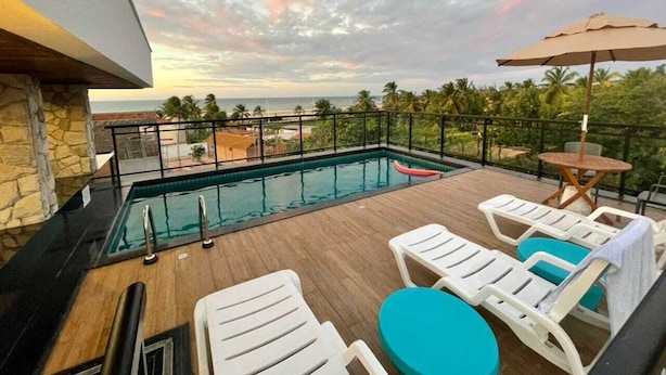 A rooftop pool area with lounge chairs and a table, surrounded by a stone wall and a view of the ocean. - Terraço Flecheiras (Trairi)