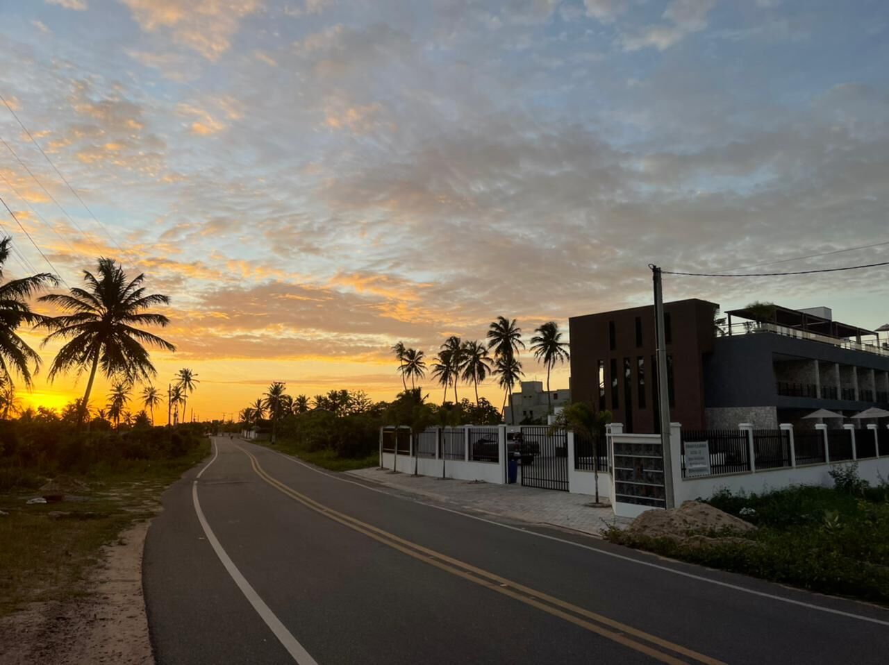 A road with a modern building on the right, palm trees, and a sunset sky.