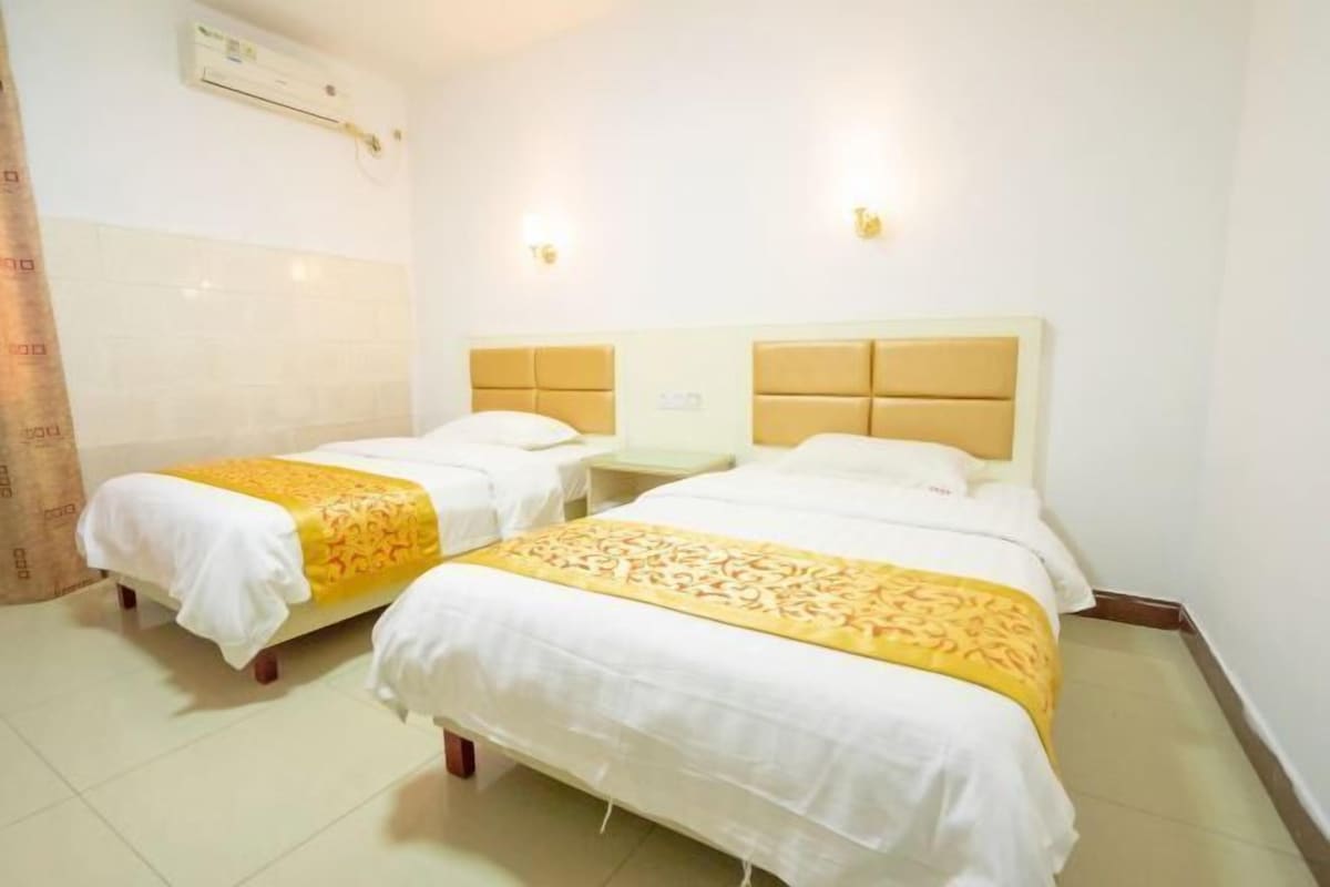 A hotel room with two beds, each with a white bedspread and a yellow decorative runner. There are two bedside tables, a wall-mounted air conditioning unit, and two wall-mounted light fixtures.