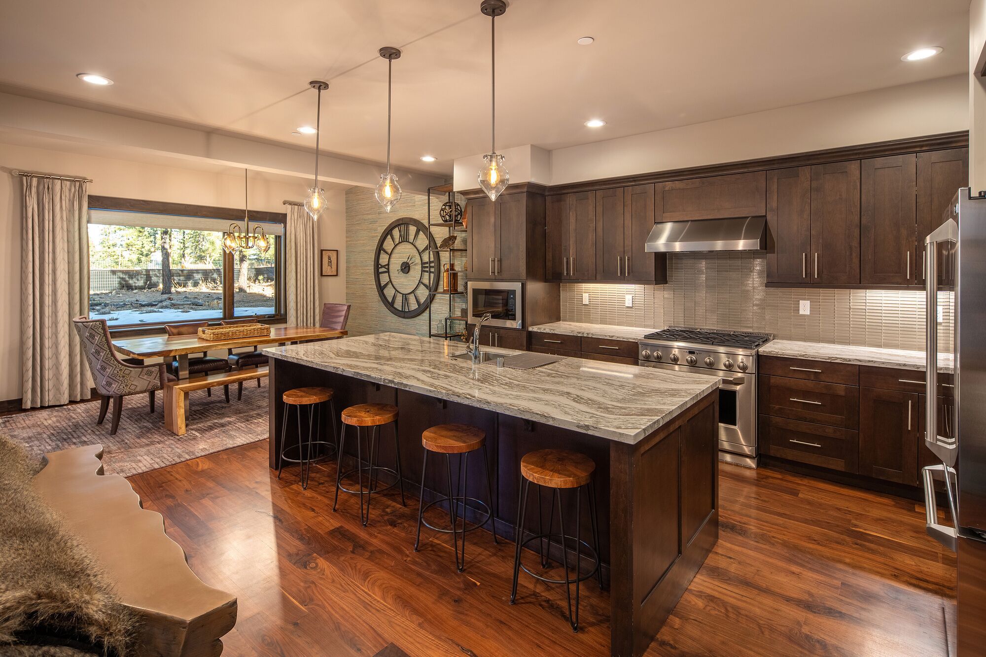 A modern kitchen with dark wood cabinets, a large island with stools, stainless steel appliances, and a large wall clock.