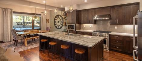 A modern kitchen with dark wood cabinets, a large island with stools, stainless steel appliances, and a large wall clock.