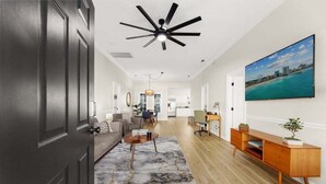 A modern living room with a ceiling fan, a large TV, a wooden console table, and a cityscape painting. - Spacious 4BR Beach House w/ Pool | Pura Vida (North Myrtle Beach)
