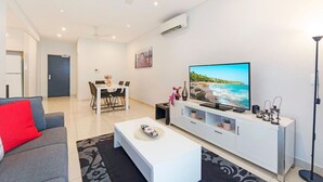 Een moderne woonkamer met een flatscreen televisie, een witte tv-kast, een grijze bank en een eethoek met een tafel en stoelen.