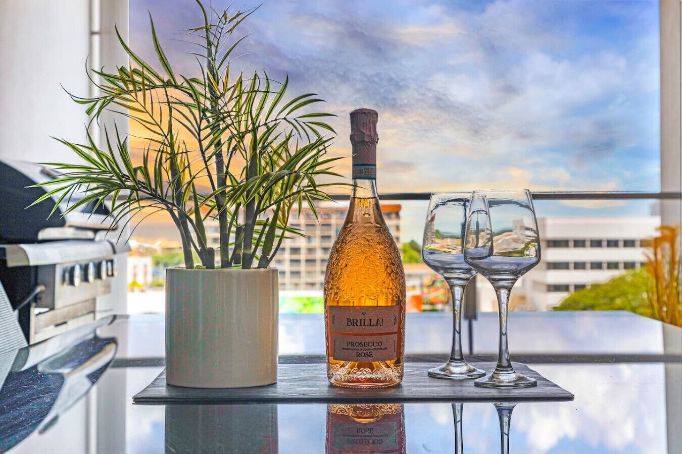 A bottle of Brilla! Prosecco Rose on a glass table with two wine glasses, a potted plant, and a view of buildings through a window.