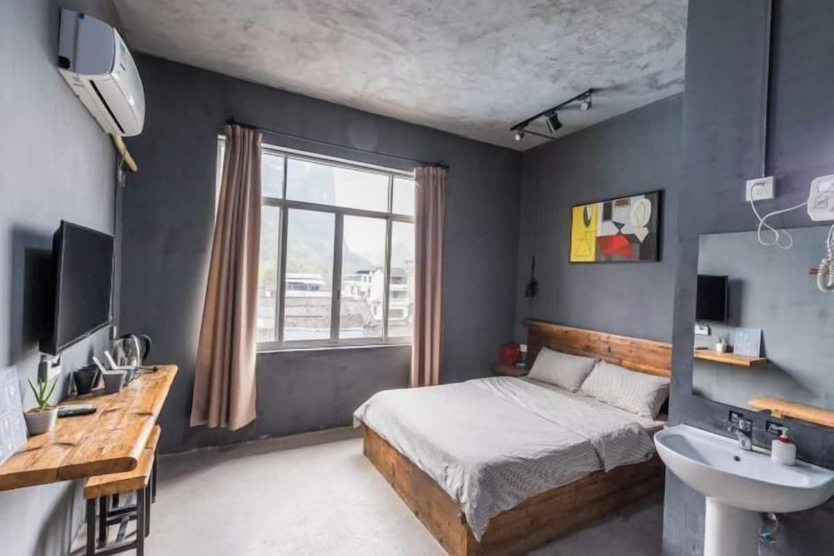 A modern bedroom with a wooden bed, a desk with a chair, a large window with curtains, a mounted TV, and a bathroom with a sink and mirror.