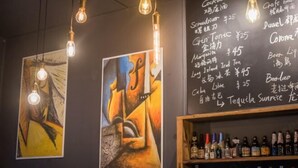 Eine Barbereich mit einer Kreidetafel, die Getränke und Preise auflistet, Kunstwerken an den Wänden und hängenden Glühbirnen.
