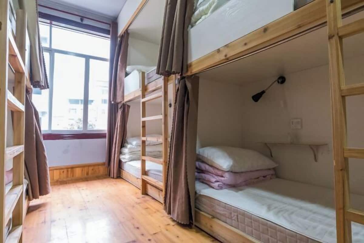 A dormitory room with bunk beds, wooden flooring, and a window with curtains.