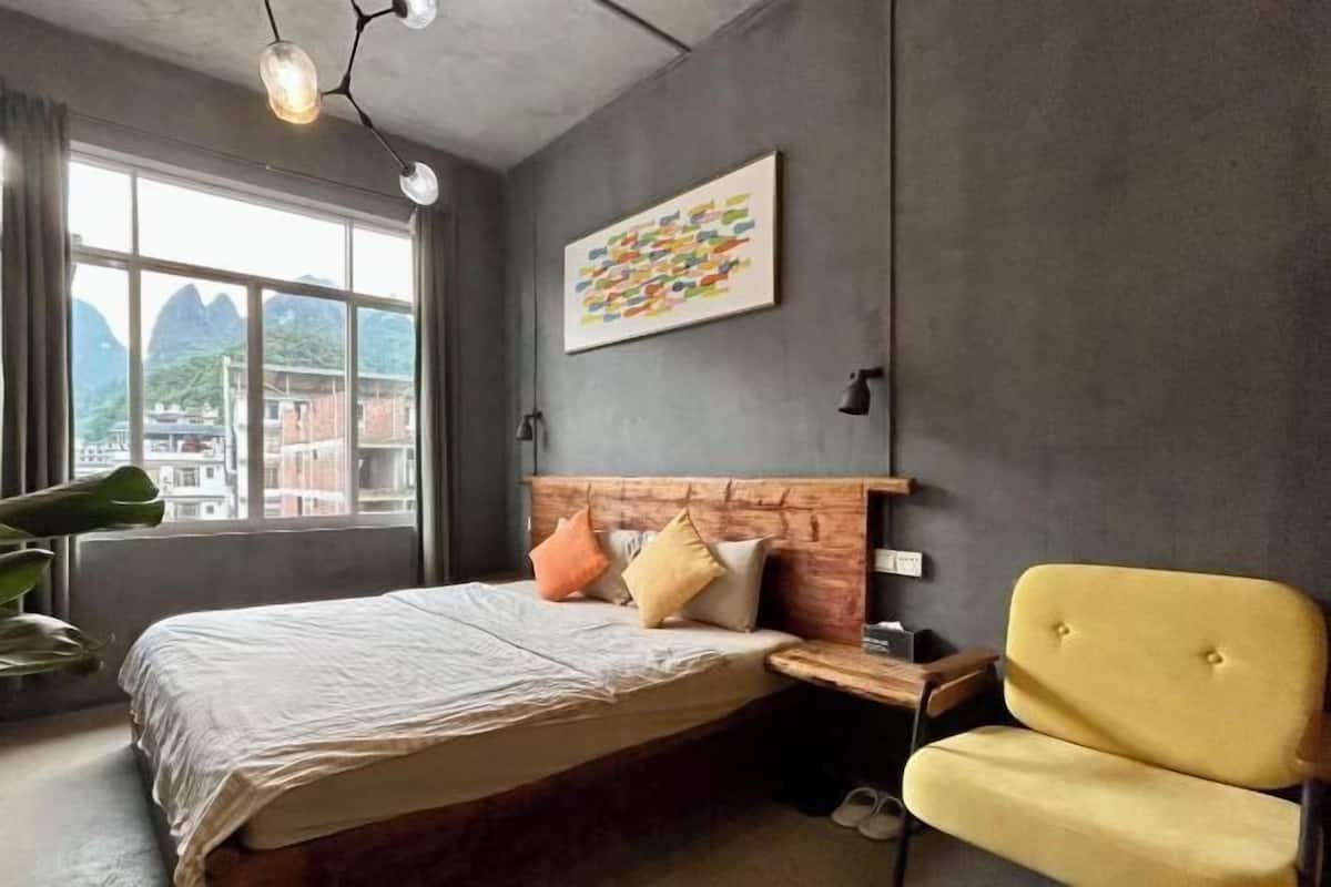 A modern bedroom with a large bed, a wooden headboard, a yellow armchair, and a view of mountains through a large window.