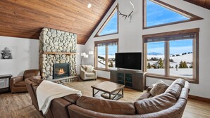 Amplia sala de estar con chimenea de piedra, sofás de cuero y grandes ventanales con vista a una montaña nevada.