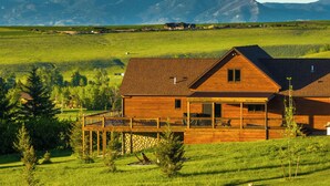 Una cabaña de madera con un porche, rodeada de campos verdes y montañas al fondo.