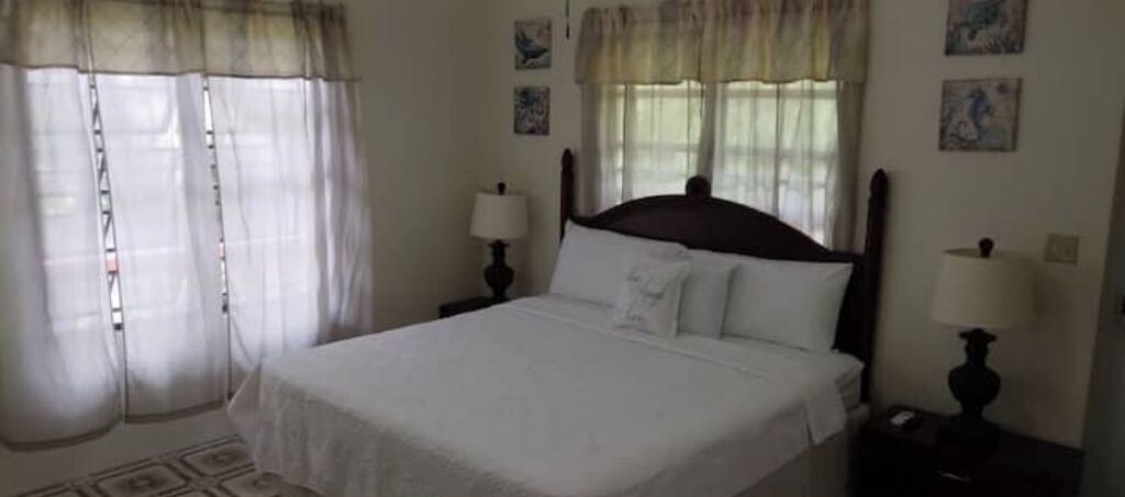 A neatly made bed with white linens, two bedside lamps, and a window with sheer curtains.