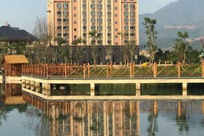 A high-rise hotel with multiple floors, a water body in front, and a wooden bridge. - Menluo Wa Jing Luxury Grand Hotel(Hulu Town Wa Tribe Branch) (Cangyuan)