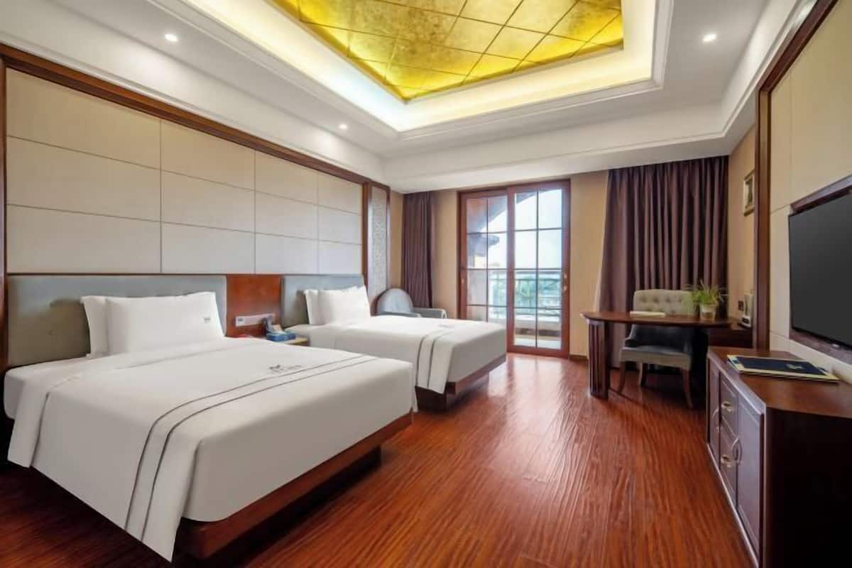 A hotel room with two beds, a desk, a television, and a balcony with curtains.