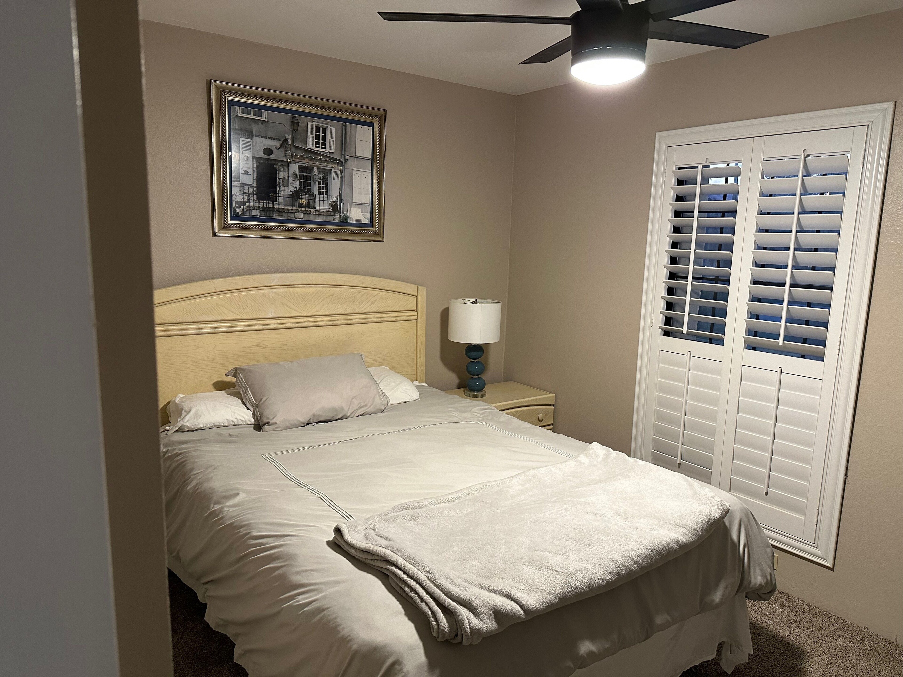 A neatly made bed with a headboard, a bedside lamp, and a window with blinds.