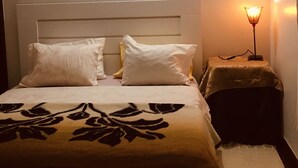 Ein Bett mit weißem Kopfteil, zwei weiße Kissen und einer decke mit Blumenmuster. Neben dem Bett steht ein kleiner Tisch mit einer Lampe.