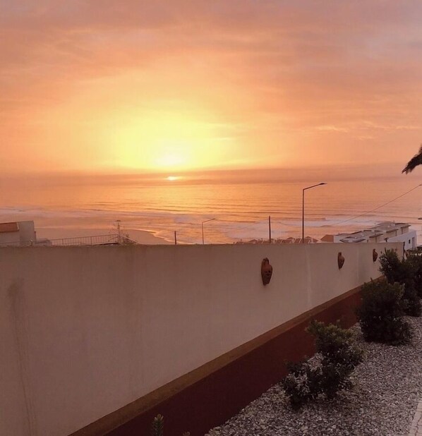 Une vue sur le coucher de soleil depuis un balcon donnant sur une plage, avec un petit muret et des lampadaires.