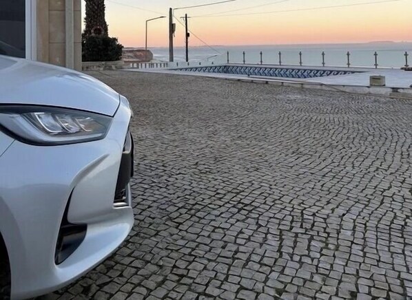 Un coche blanco aparcado en una calle empedrada, con vista al mar y un paseo marítimo al fondo.