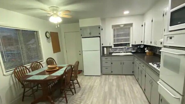 A compact kitchen with white cabinets, a dining table with chairs, a ceiling fan, and a window with blinds. - The Little Fish in Eufaula (Eufaula)