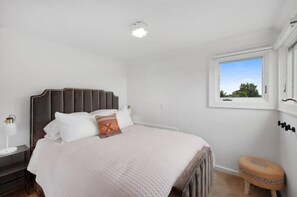 A bedroom with a bed, a nightstand, a lamp, a window, and a small stool.