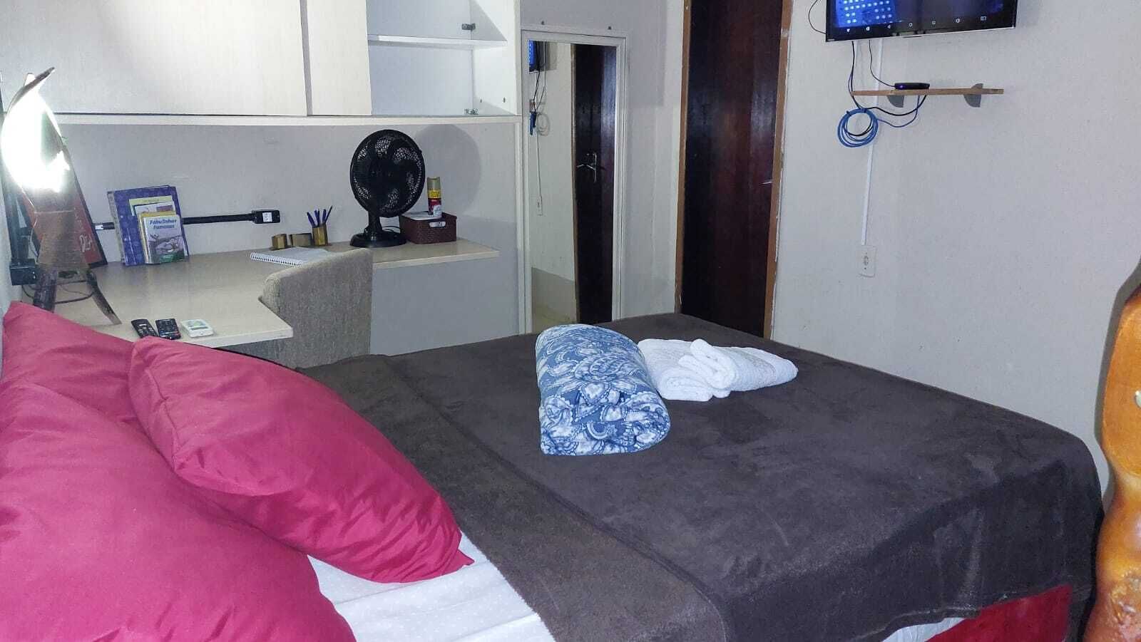 A bedroom with a bed, pillows, a fan, a TV, and a desk with a computer and some items on it.