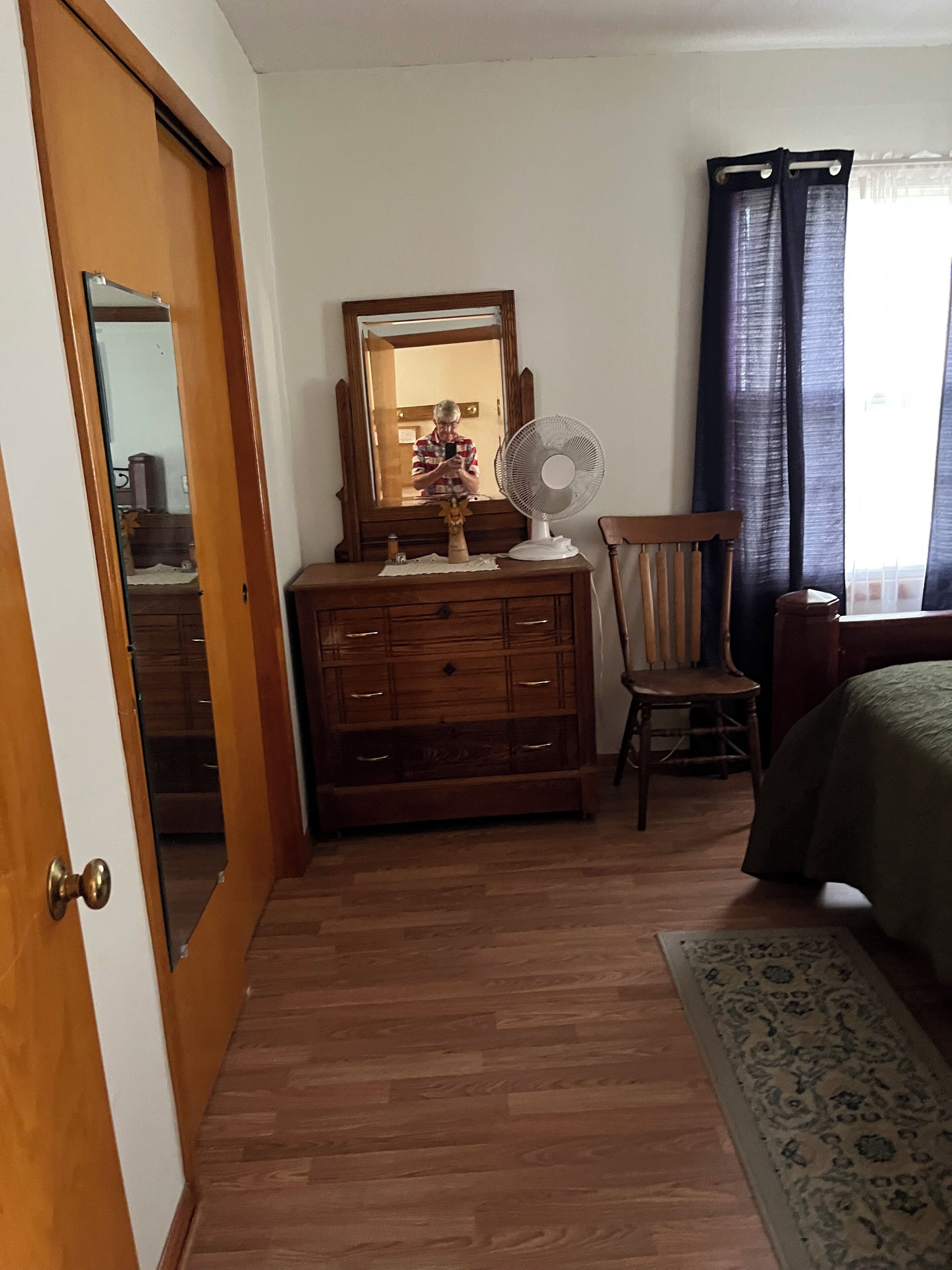 Quarto com cômoda de madeira, uma cadeira, uma cama, um espelho e uma janela com cortinas.