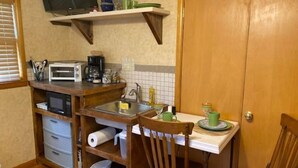 A compact kitchen with a microwave, toaster, and coffee maker on a wooden countertop. There is a sink with a faucet, a chair, and a small table with a green cup and saucer. A wooden door is visible in the background. - Welcome to our Wichita Home - Easy Close Access to Turnpike South Side (Wichita)
