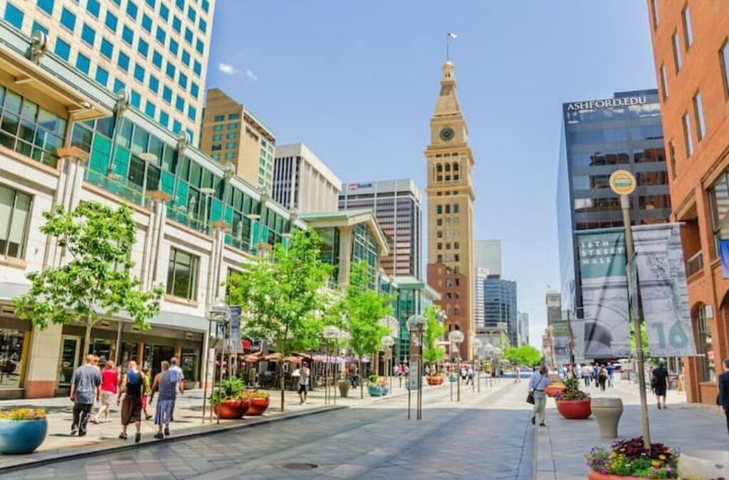 A city street with modern buildings, a prominent clock tower, and pedestrians.