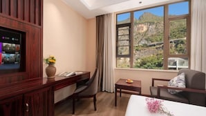 A hotel room with a desk, chair, bed, TV, and a window with a mountain view. - Fall In Love With Nujiang Grand Canyon Hotel (Lushui)