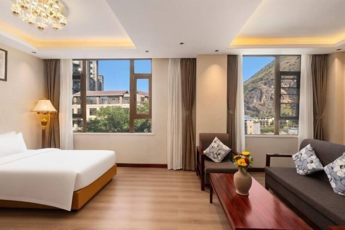 Ein Hotelzimmer mit Bett, einer Sitzecke, einem Tisch mit Blumenvase und großen Fenstern mit Blick auf die Berge.