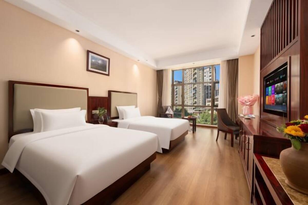 Ein Hotelzimmer mit zwei Betten, einem Schreibtisch, einem Fernseher und einem großen Fenster mit Blick auf die Stadt.