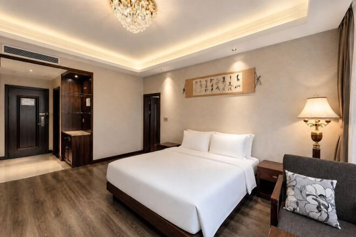 A hotel room with a large bed, a seating area with a chair and a table, a wall-mounted artwork, and a chandelier.