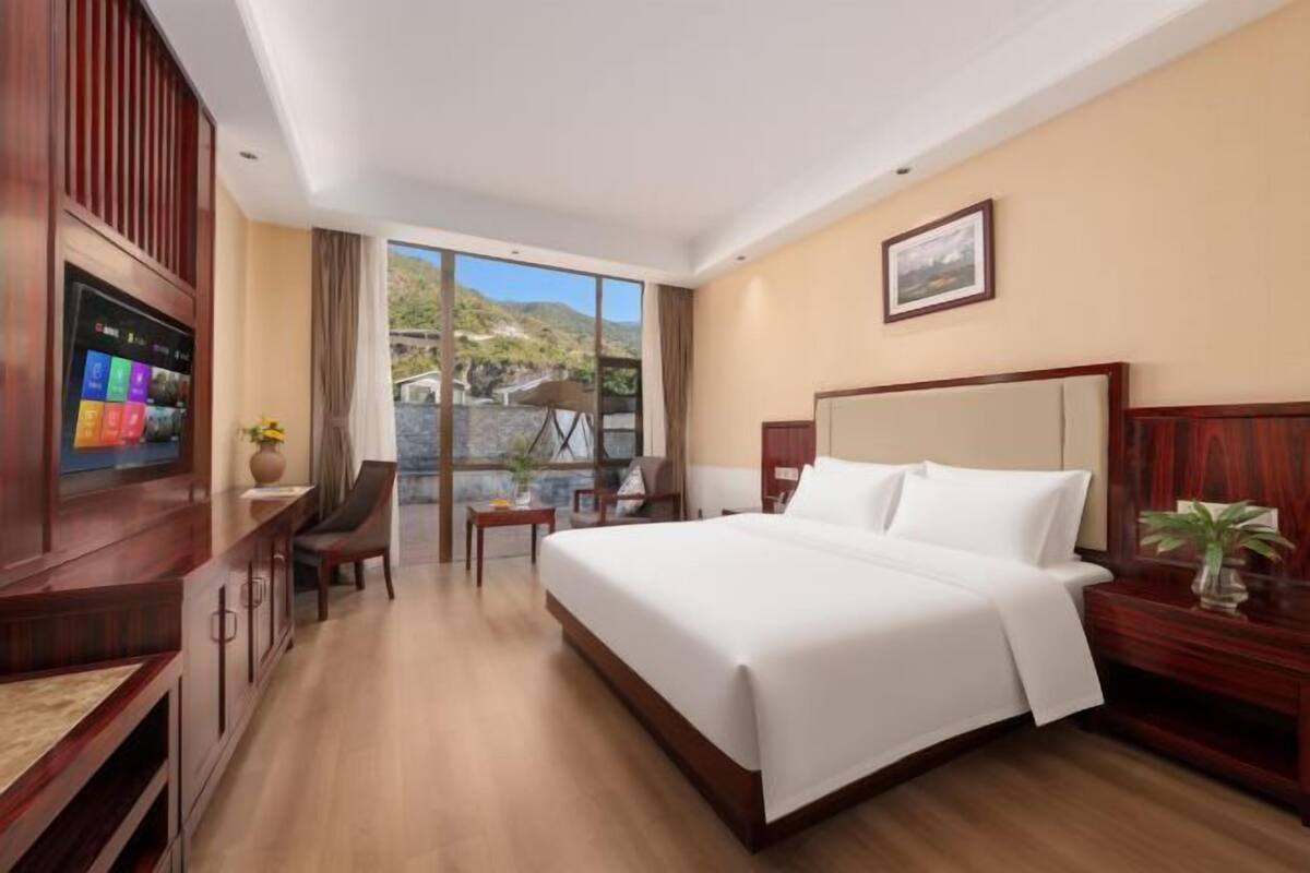 Ein Hotelzimmer mit einem großen Bett, einem Fernseher, einem Schreibtisch und einem Fenster mit Blick auf Hügel.