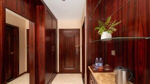 Un couloir avec des portes en bois, une étagère en verre sur laquelle est posée une plante, un plan de travail avec une bouilloire, et une porte au fond.