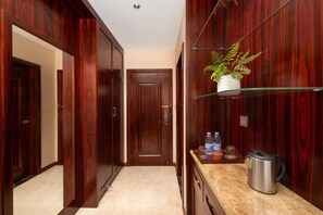 Un couloir avec des portes en bois, une étagère en verre sur laquelle est posée une plante, un plan de travail avec une bouilloire, et une porte au fond.