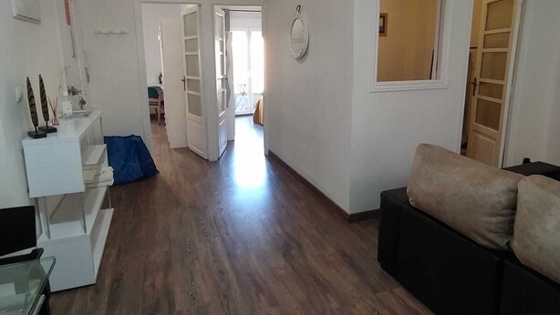Een ruime woonkamer met een houten vloer, een zwarte bank en een witte kast. Er is een deur naar een andere kamer en een raam met uitzicht.