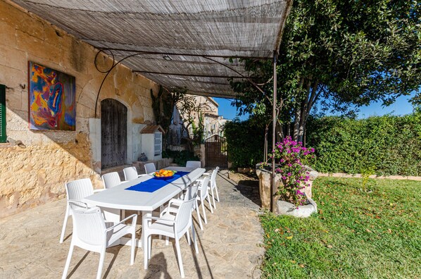 An outdoor dining area with a table and chairs, a stone wall, and a garden with a tree and flowers. - YourHouse Can Bombarda, large house with private pool (Llubi)