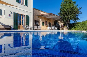 A swimming pool with a tiled edge, a covered outdoor seating area with chairs, and a building with a stone facade and green shutters. - YourHouse Can Bombarda, large house with private pool (Llubi)