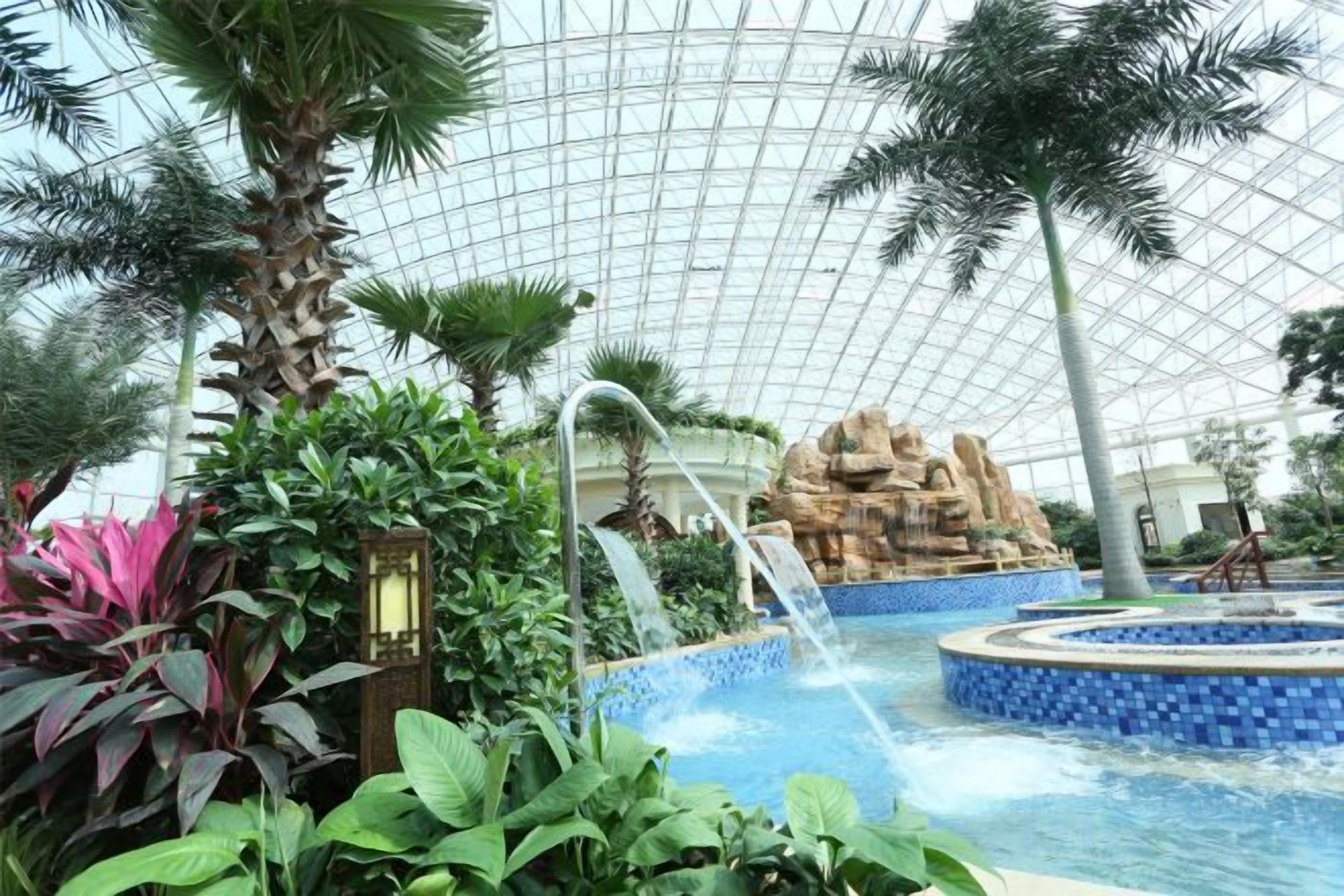 An indoor pool area with a waterfall feature, surrounded by tropical plants and a glass ceiling.