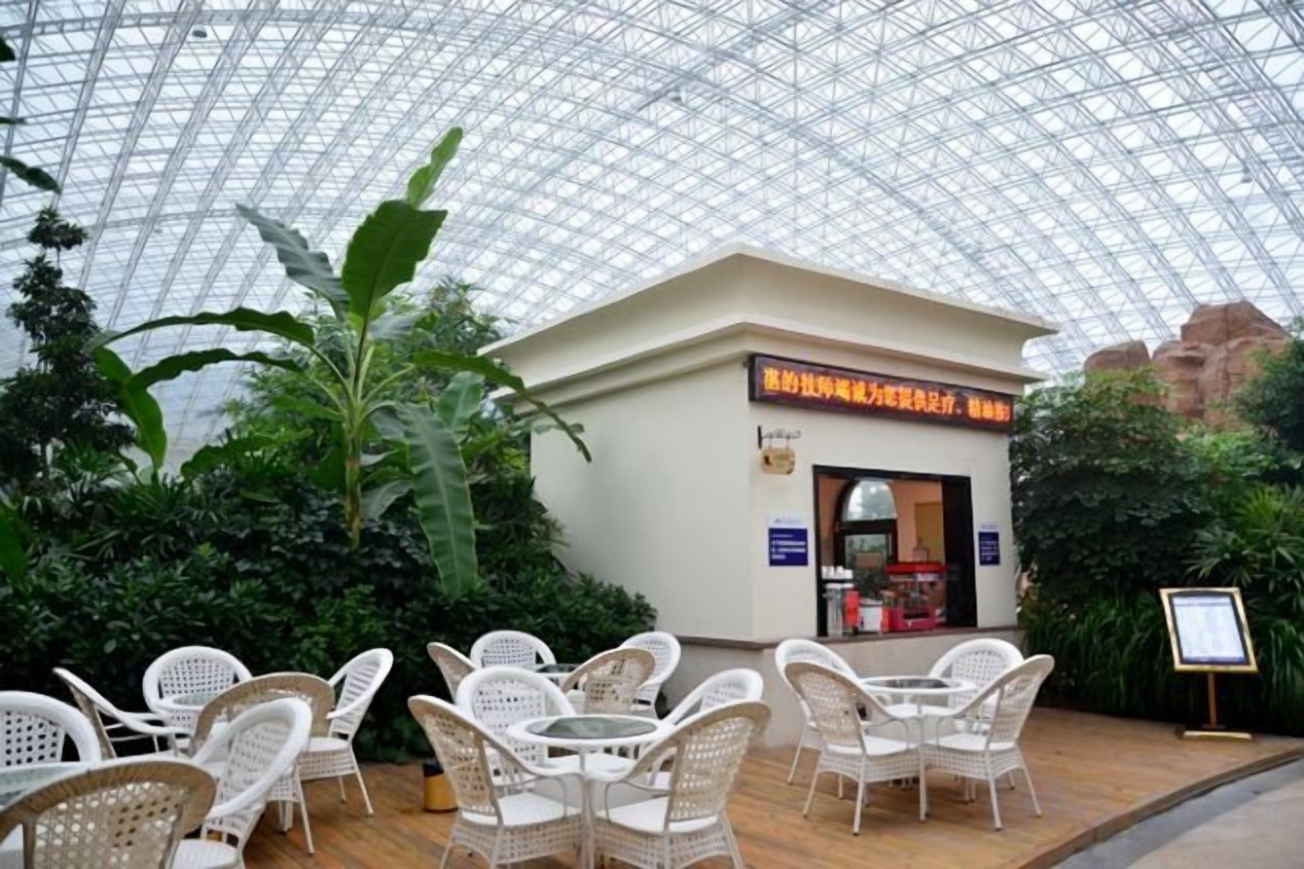 An indoor restaurant with a glass ceiling, surrounded by greenery and white wicker furniture.
