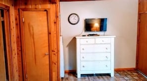 Una habitación con una puerta de madera, un aparador blanco, un televisor y un reloj de pared.