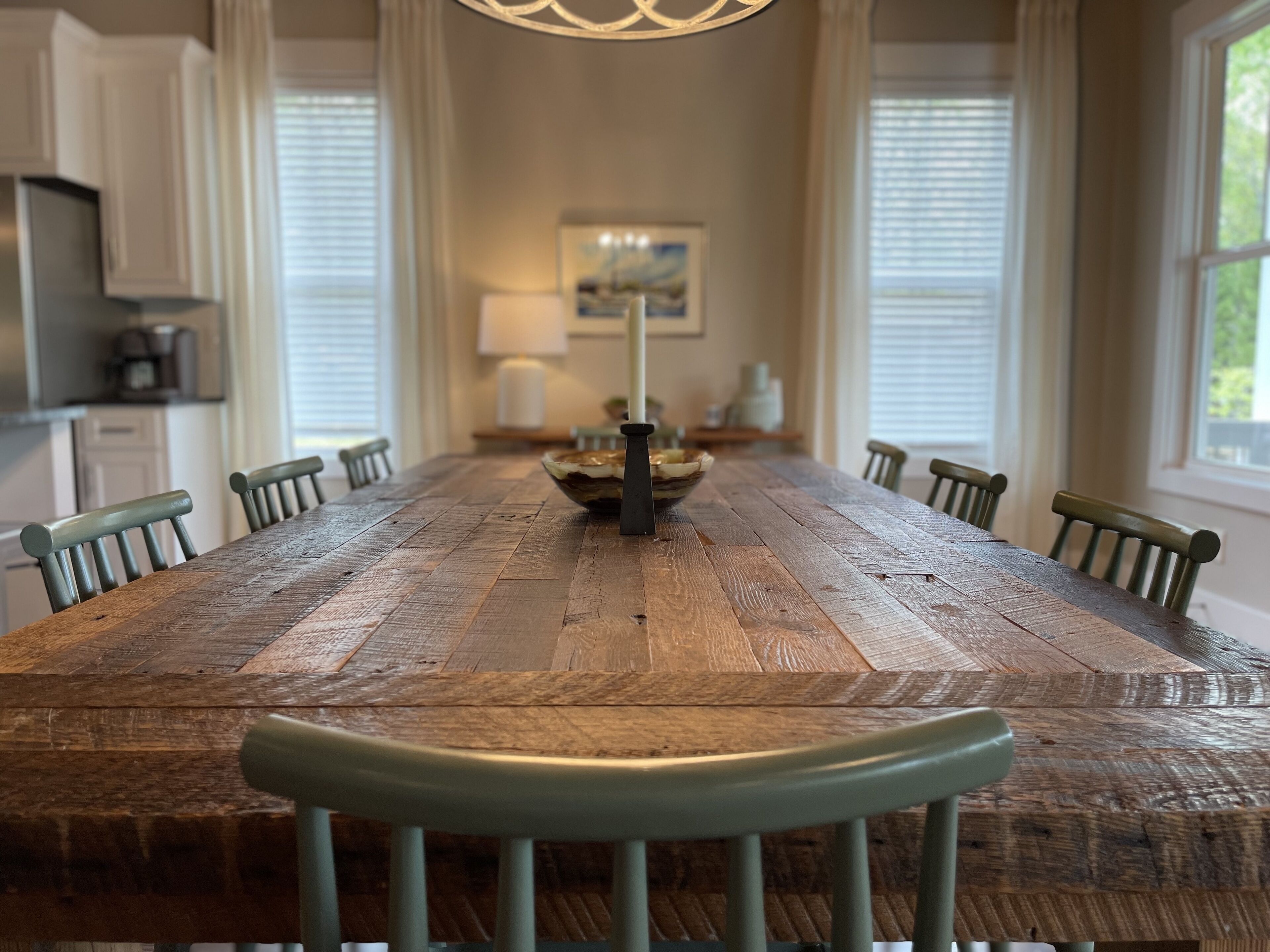 Une longue table à manger en bois, entourée de chaises, un lustre et, au fond, une vue sur la cuisine.