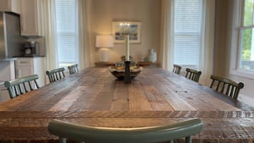 Une longue table à manger en bois, entourée de chaises, un lustre et, au fond, une vue sur la cuisine.