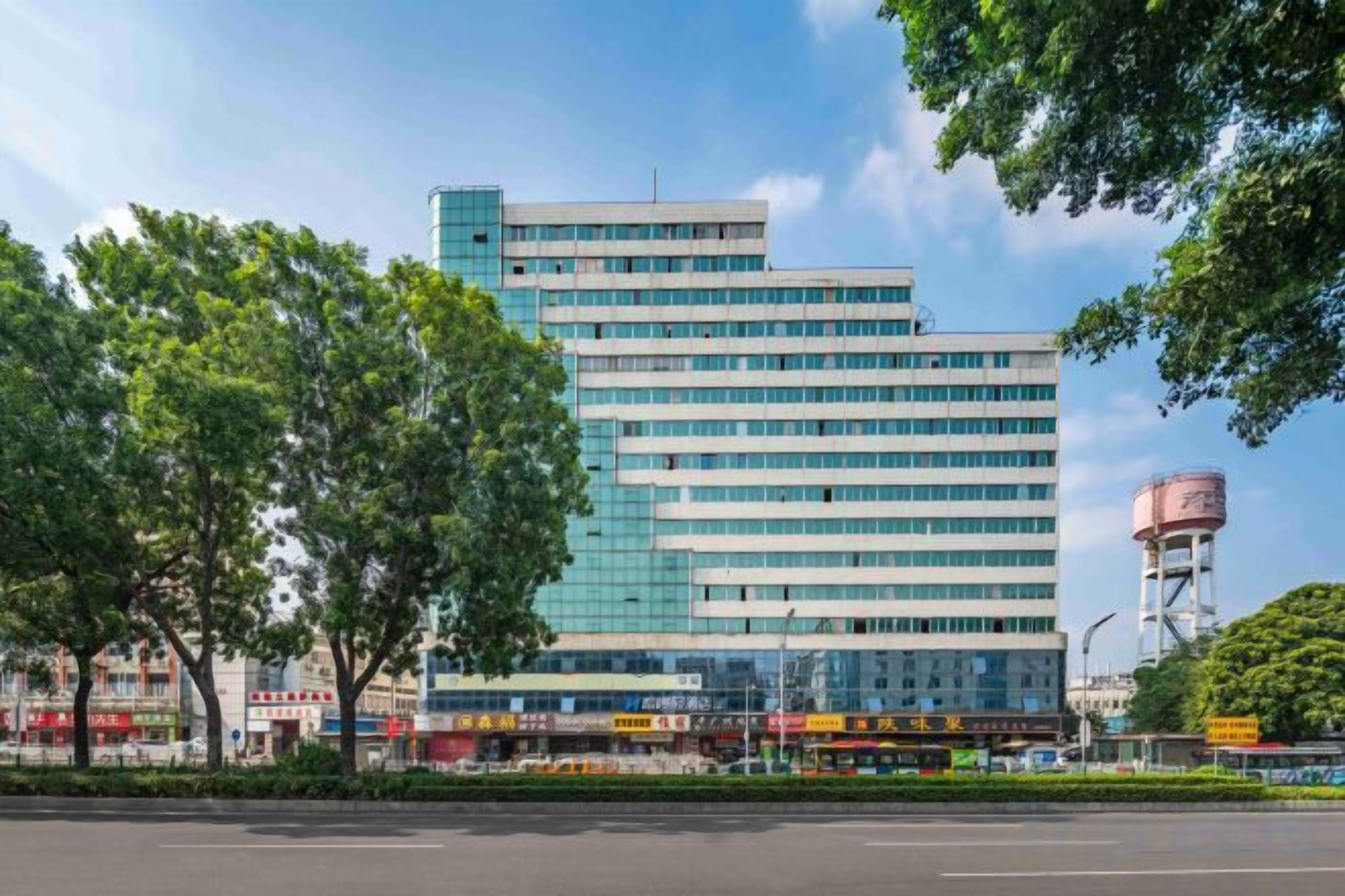 Un edificio de varios pisos con fachada de vidrio, rodeado de árboles y una torre de agua al fondo.