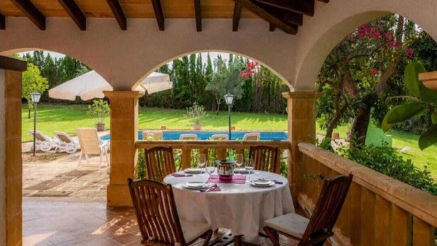 Eine überdachte Terrasse mit Esstisch, mit Blick auf einen Swimmingpool und einen üppigen Garten.