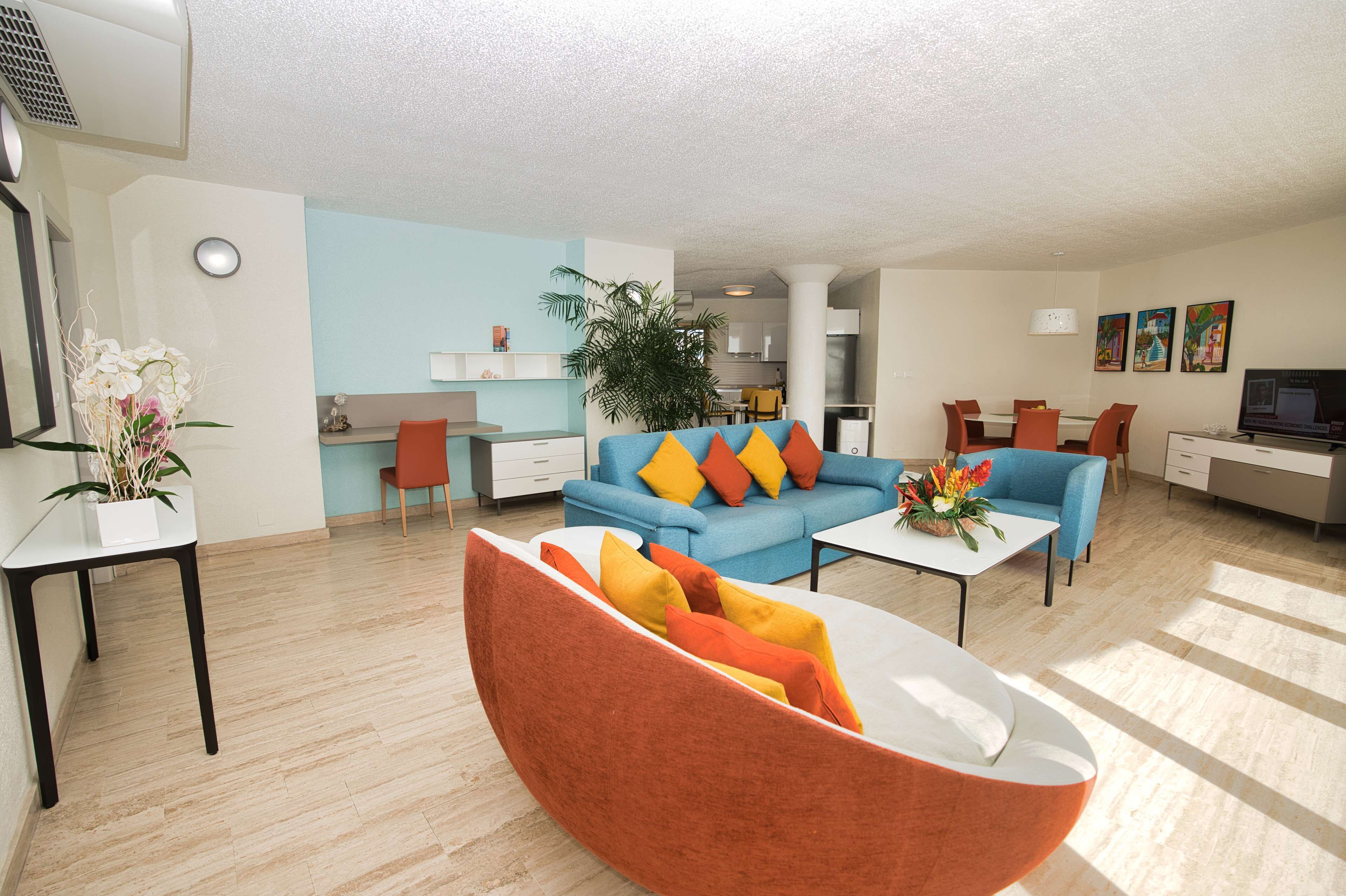 Un salon moderne avec un canapé bleu, un fauteuil orange et un coin repas comprenant une table et des chaises blanches.