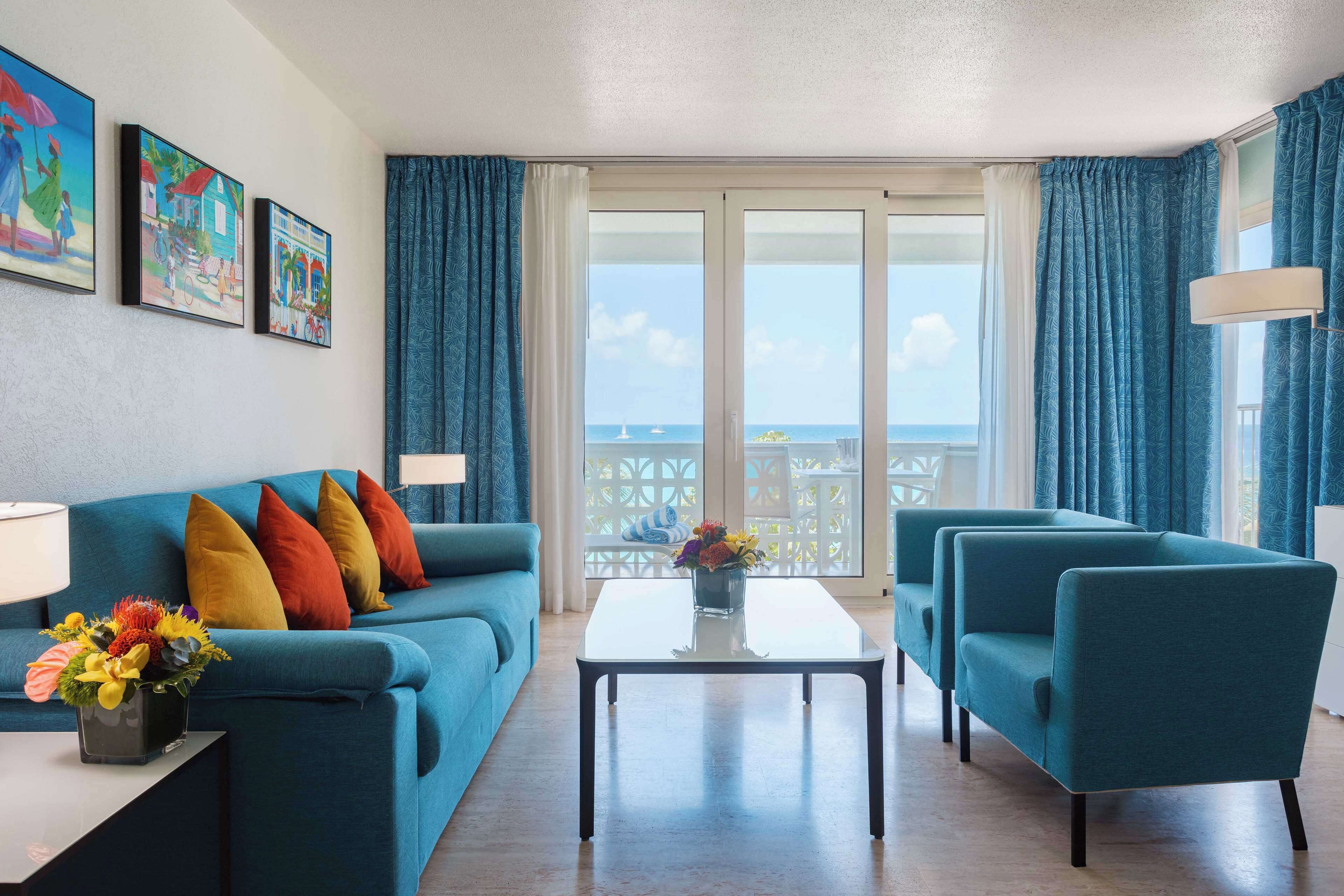A modern living room with a blue sofa, matching armchairs, and a coffee table. There are three framed artworks on the wall, and a balcony with a view of the sea.