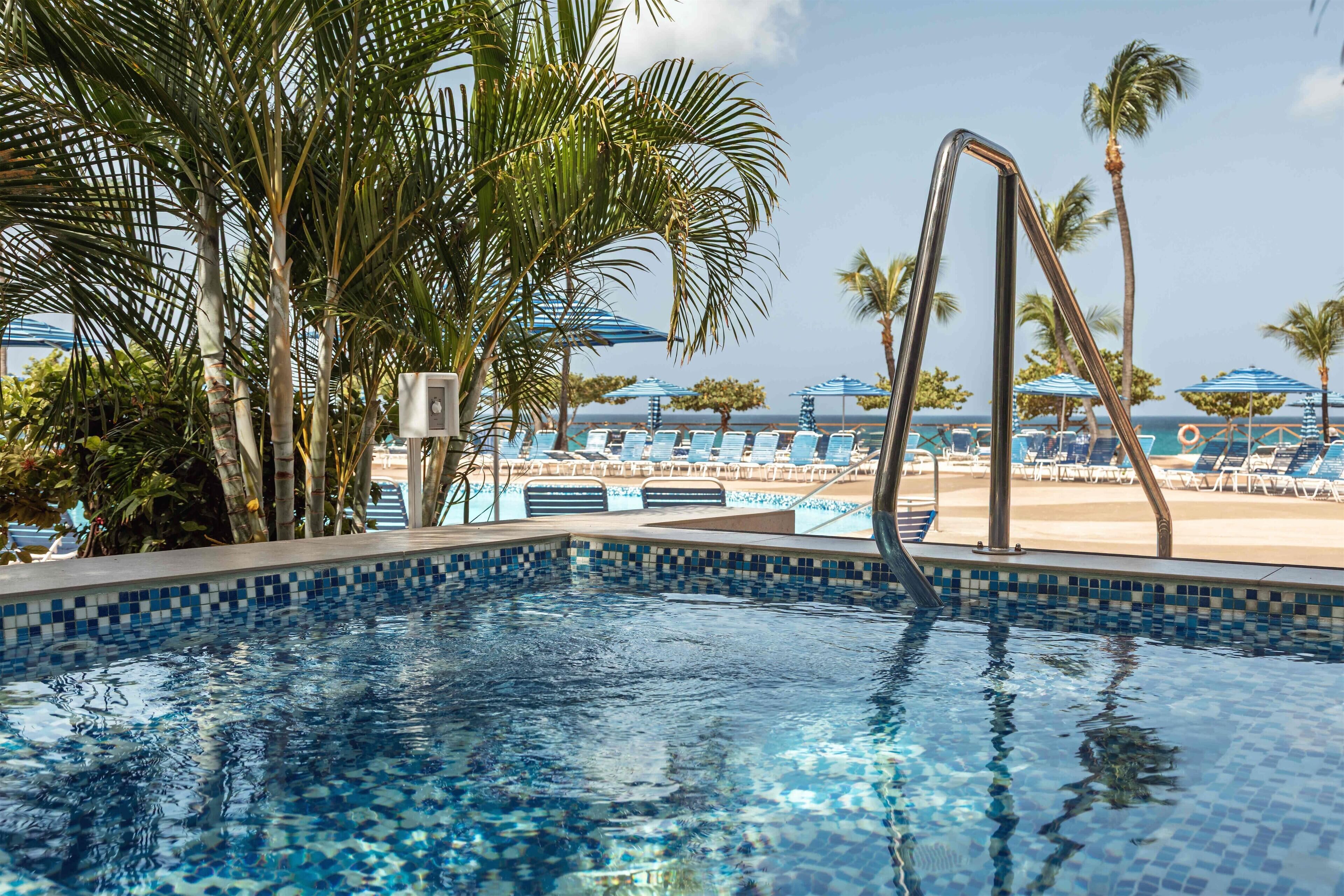 A swimming pool with clear blue water, surrounded by lounge chairs and palm trees, with a view of the beach and ocean in the background.